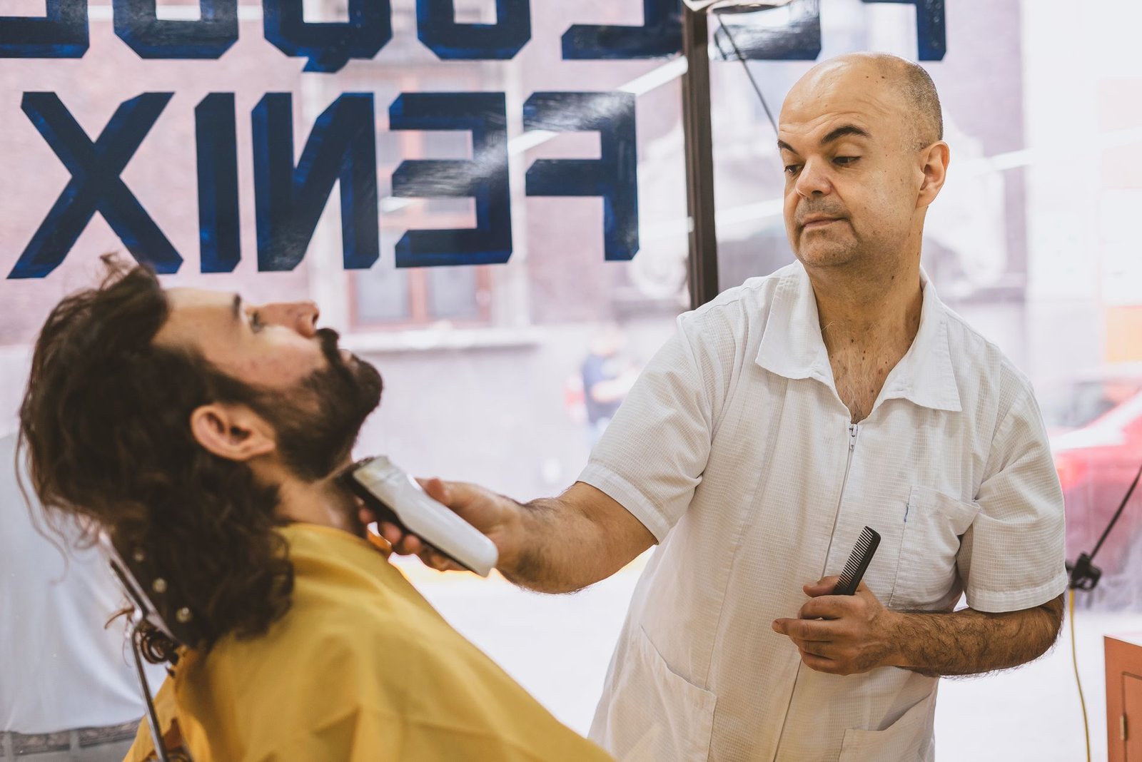 Barbeiro fazendo a barba de um cliente com máquina em uma barbearia de bairro